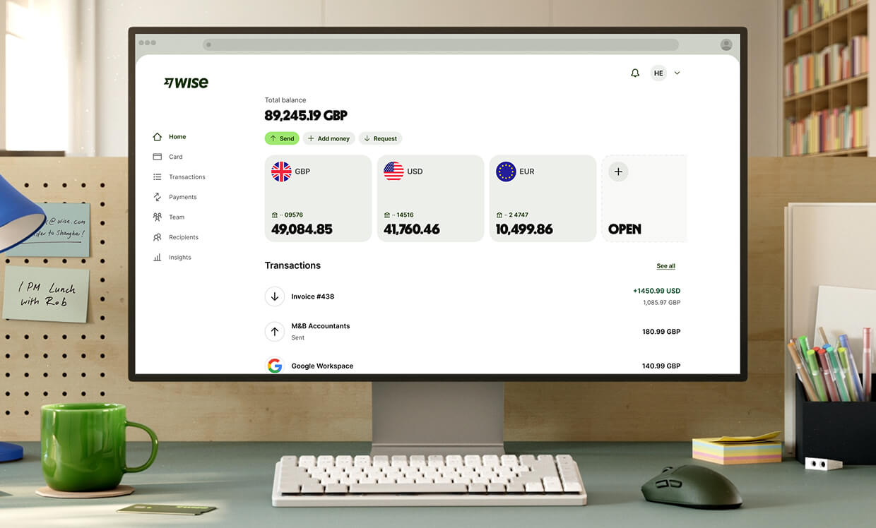 A close-up shot of a modern workspace shows a white computer monitor displaying the Wise dashboard. The screen shows a total balance of "89,245.19 GBP" and three main currency accounts: GBP with "49,084.85," USD with "41,760.46," and EUR with "10,499.86," followed by an option to open a new account. Recent transactions are listed below. In front of the monitor is a compact white keyboard, a dark grey mouse, and a bright green ceramic mug.