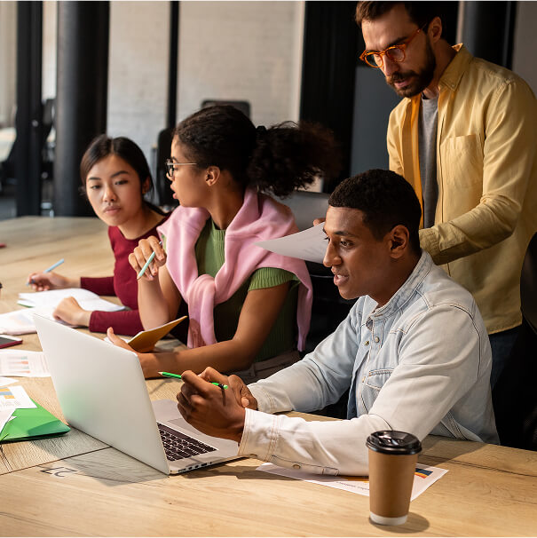 A diverse group of four individuals is gathered around a conference table. In the foreground, a man sits at the table, looking at an open laptop and holding a pen. To his left, another man in a yellow shirt and leans over, looking at the laptop screen. Behind the seated man, two women are visible; one is writing, and the other is looking off to the side. There is a changing banner that shows different amounts of money in different currencies.
