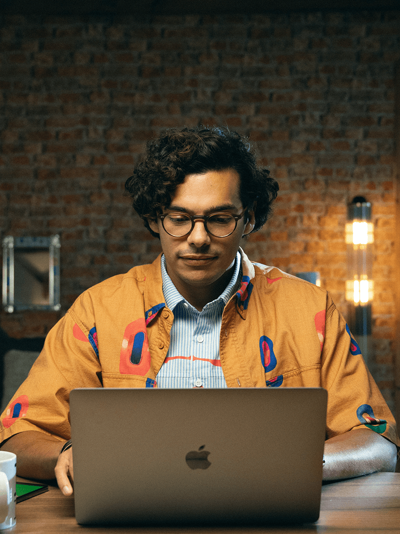 A man with glasses sits at a desk, looking down at a laptop. He is wearing a distinctive orange jacket. In the background, a brick wall is visible. The words "FOR SMALL BUSINESSES" are displayed in white text at the top of the image, and a green circle with a white plus sign is in the bottom right corner.