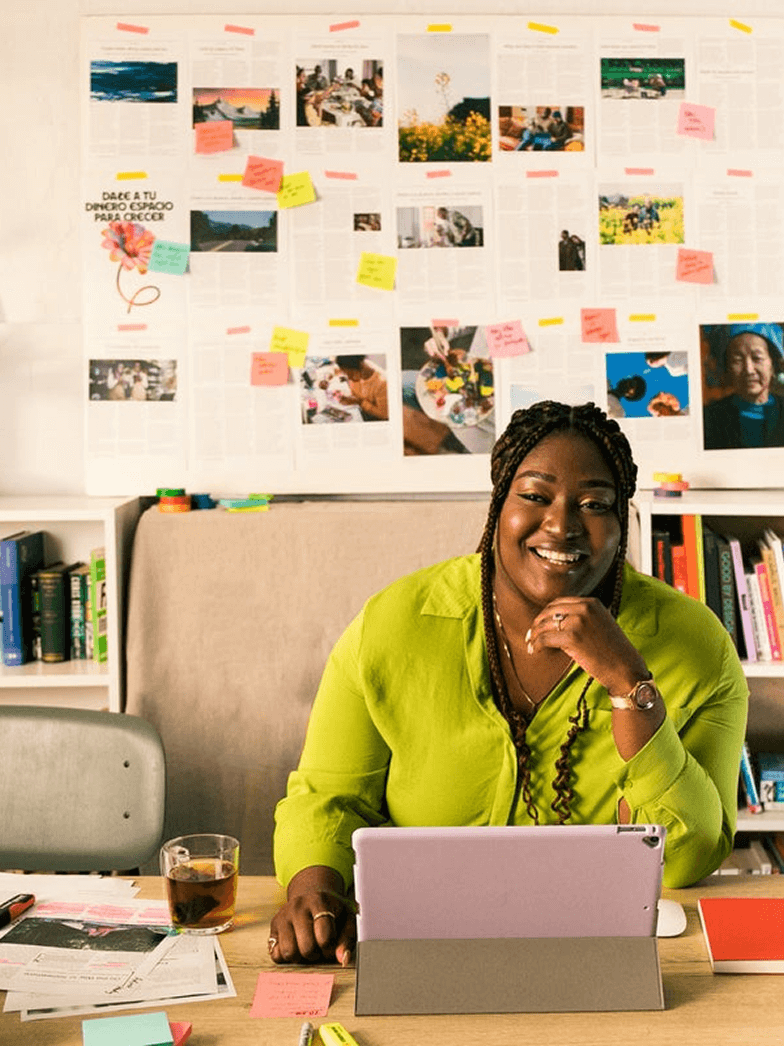 A woman wearing a green shirt sits at a desk, smiling. In front of them is a purple tablet propped up in a grey case. Behind them, a wall is covered with various photos, drawings, and numerous colourful sticky notes, suggesting a creative workspace. The words "FOR STARTUPS" are displayed in white text at the top of the image, and a green circle with a white plus sign is in the bottom right corner.